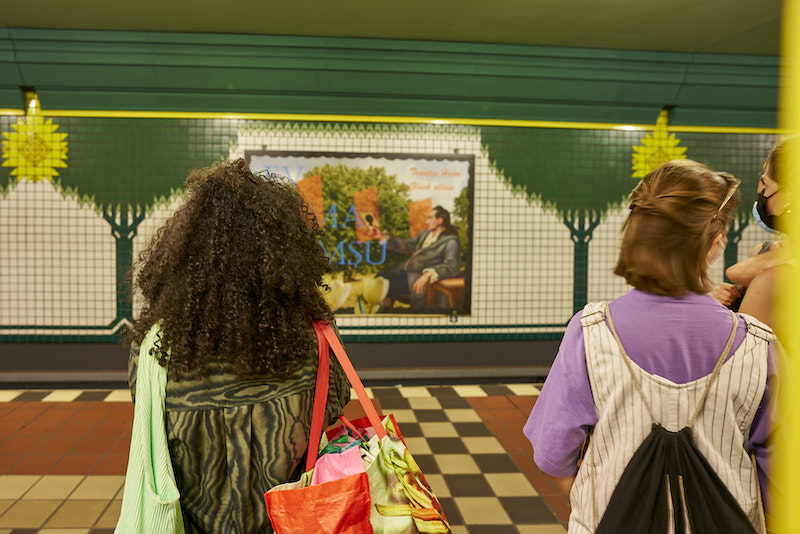 two women stood on the platform of an underground station looking at an artwork on the wall