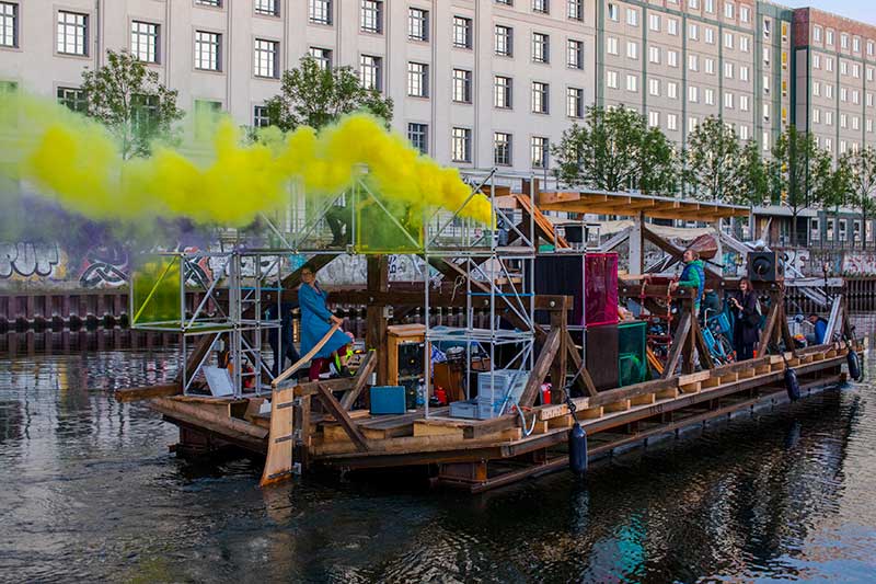 a make shift, human powered boat spewing yellow smoke on an urban river