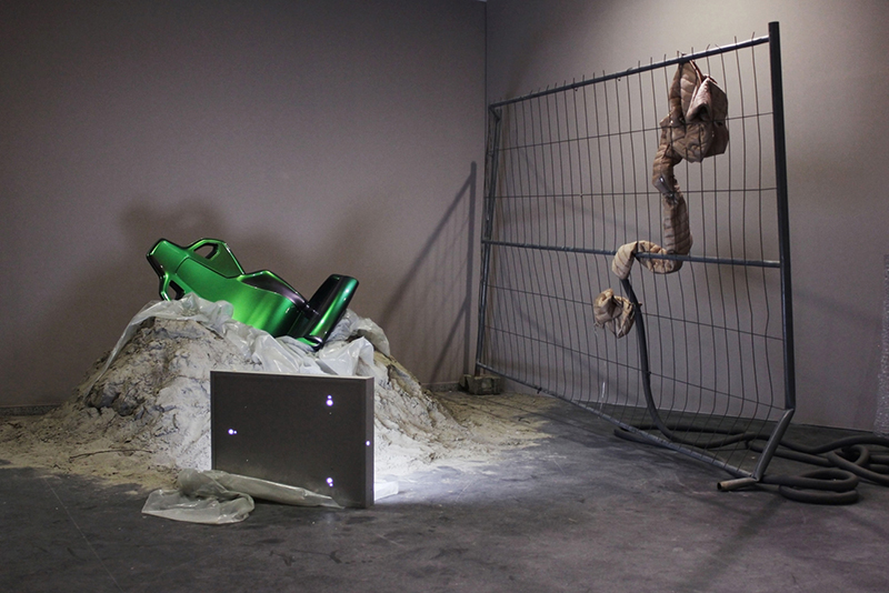 a grey room in which a green chair lies on a pile of sand next to a wire fence and a monitor