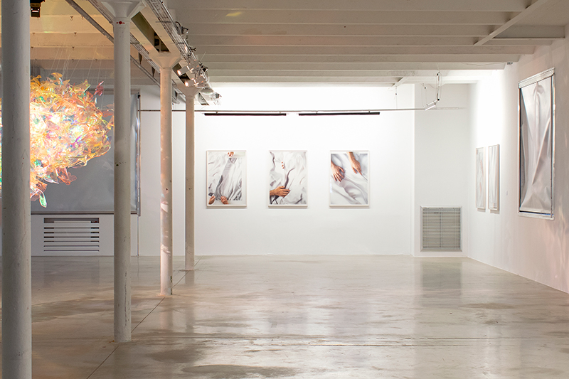 View of an exhibition with three photos hanging on a wall  and an amorphous shiny object hanging from the ceiling to the left