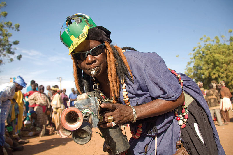 Picture of an African woman outside playing what looks like a homemade instrument with horns and whistles