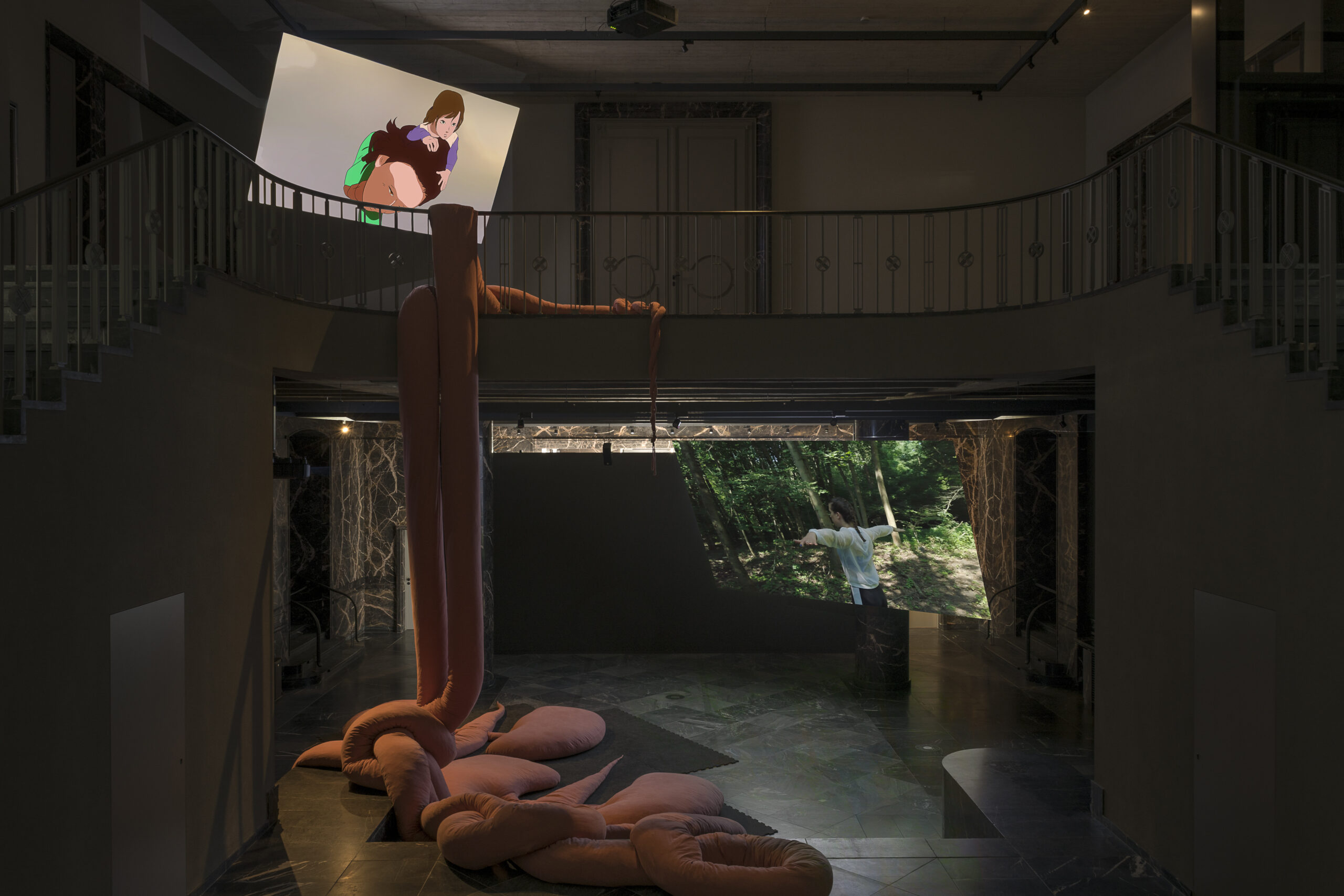 a two-storey exhibition space with large video projections and a plush tentacle-like sculpture in the middle