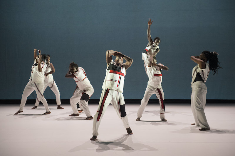 Still from a dance performance, dancers are all wearing white clothing with black and red stripes, against a cream floor and teal blue backdrop