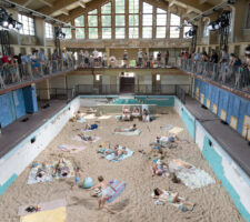 A large two storey warehouse interior with a sandy beach installed on the ground floor, with people on towels, and an audience of on-lookers on the first floor balcony along the entire inner circumference of the building