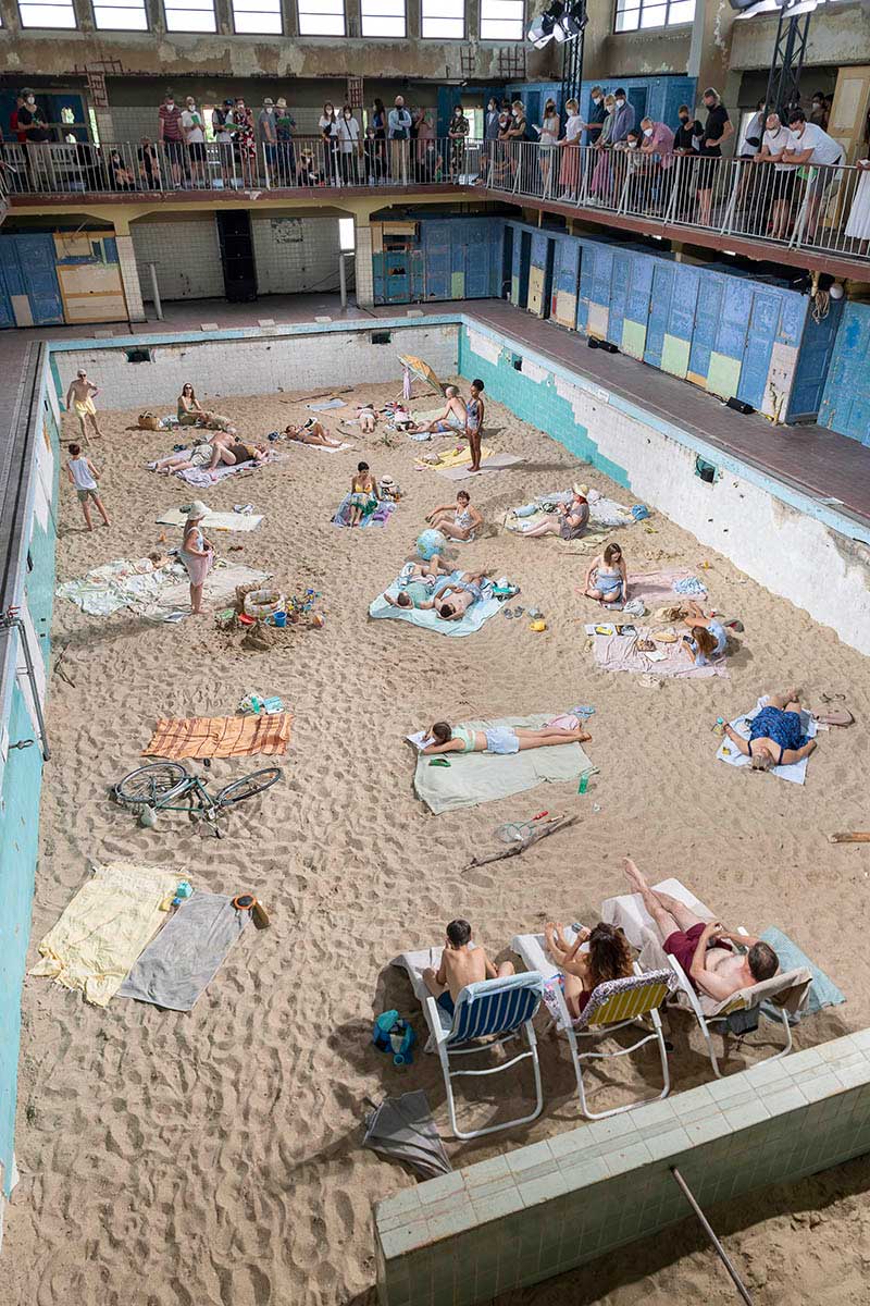a sandy beach installed at the bottom of an indoor swimming pool with people on towels lounging around