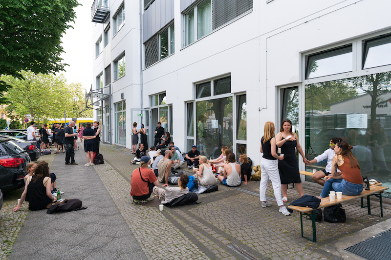 people stood around or sitting on the floor outside a building with white facades and large windows