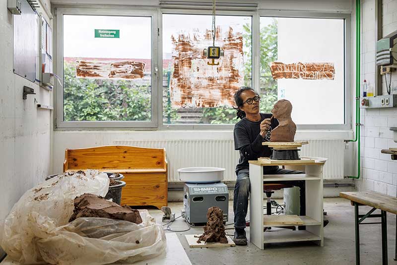 A man sits in a ceramic workshop room making a bust out of clay