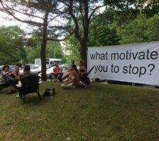 A group of people sitting in a park with a banner saying "What motivates you to stop" strung between two trees