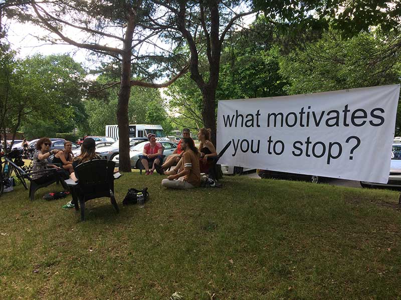 A group of people sitting in a park with a banner saying "What motivates you to stop" strung between two trees