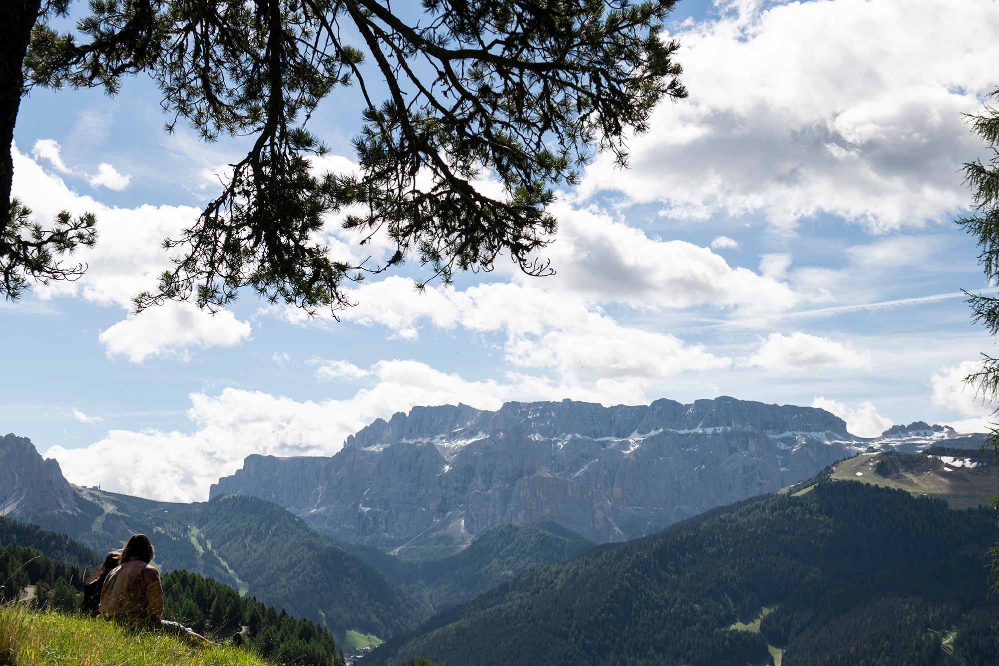 two people sitting on a hill, looking at the mountins and cloudy sky in he background