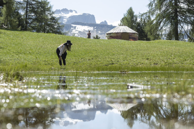 person leaning next to a pond, a yurt and mountains in the background