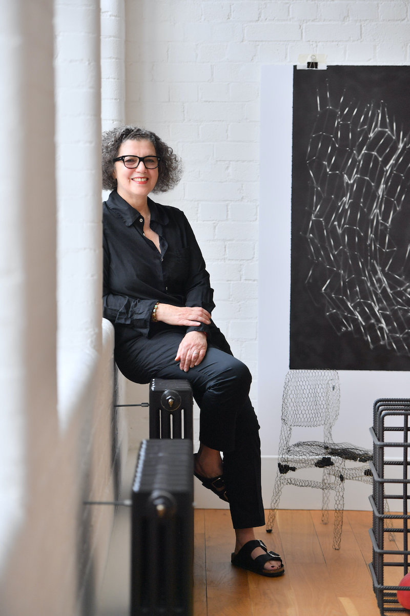 Portrait photo of Mona Hatoum, setting against a window ledge smiling, with a black painting against a white wall behind her