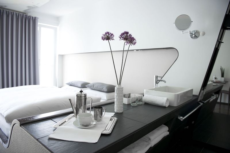 futuristic, modern design hotel room with bed, shelfs with flowers in a vase, tea pot and cup, and sink