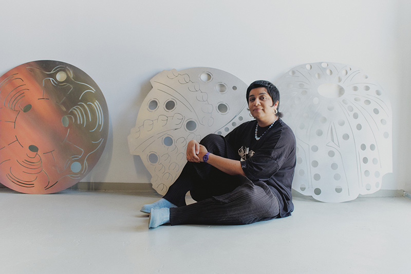 Monira Al Qadiri sitting on the floor in front of a series of aluminium sheets cut into cicles with designs in them
