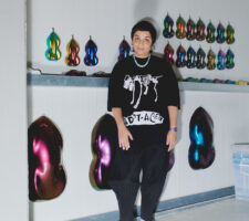 Monira Al Qadiri posing in front of a wall with iridescent plastic wall works hanging on it