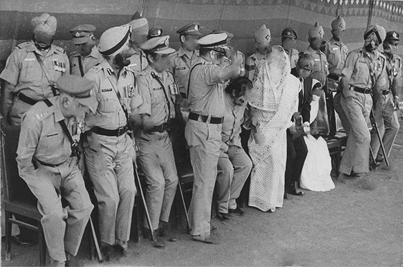 A black and white video still of Indian men in uniform and what looks like a veiled woman