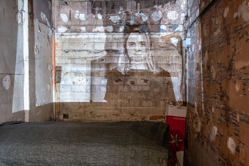 room with brick walls and a projection of a young girl in black and white is projected onto the wall straight ahead. a bed with a grey blanket sits below