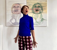 woman in a blue jumper and red checkered trousers stands in front of two drawings of faces screaming or crying. the woman is also stood screaming or crying