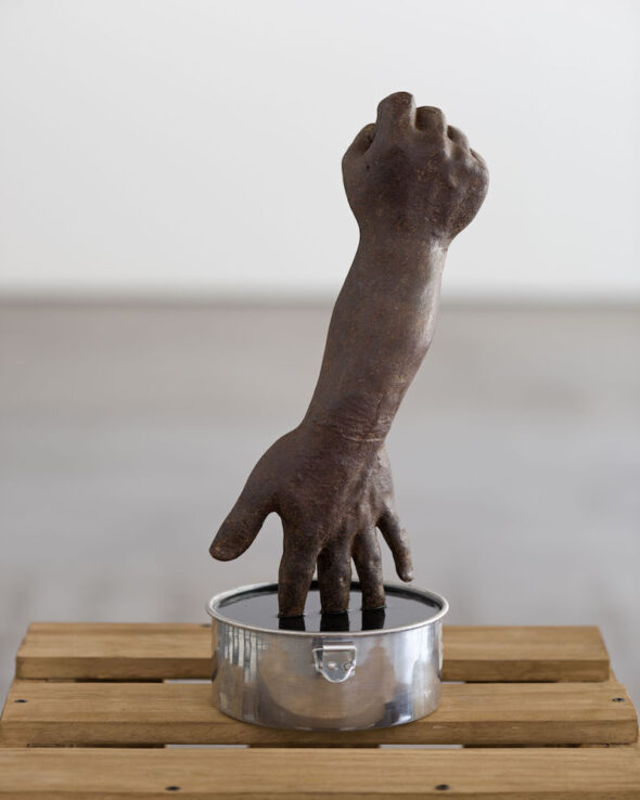 Brown sculpture of an arm with hands on both sides dipped into a liquid inside of a metal bowl
