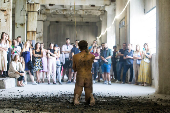 The artist is naked, kneeling on the dirt covered floor with a rope tied around his neck in front of an audience.