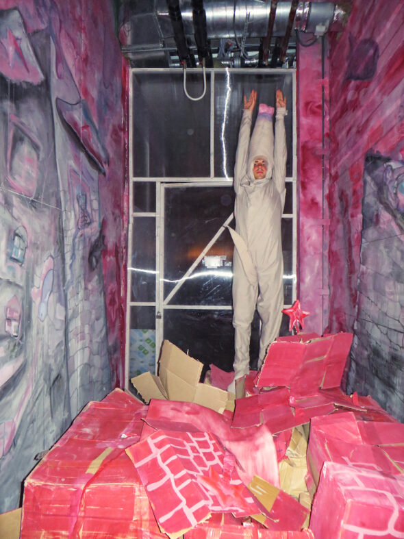 Artist in corner of small room painted with grey, pointed body suit. The walls on either side are painted with pink and grey and on the floor there are red cardboard boxes.