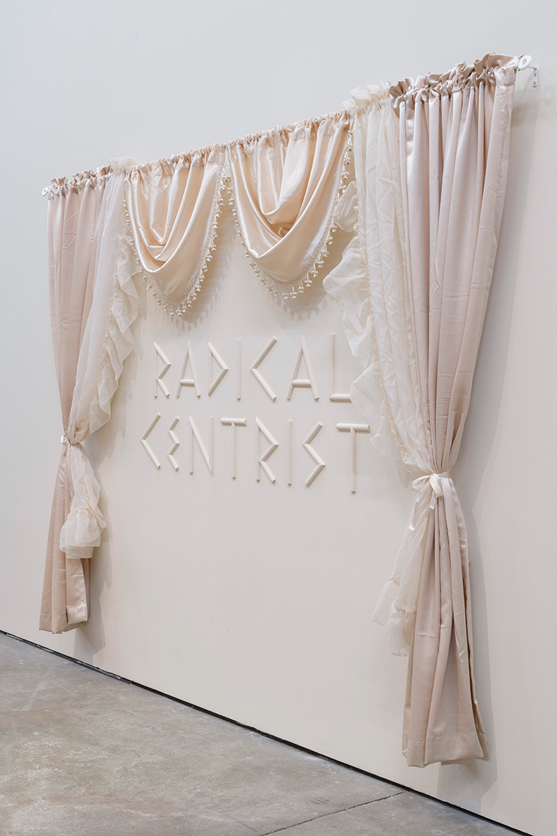 Light pink decorative curtains on a rod hanging on a white gallery wall with the words "radical centrist" embossed on the wall