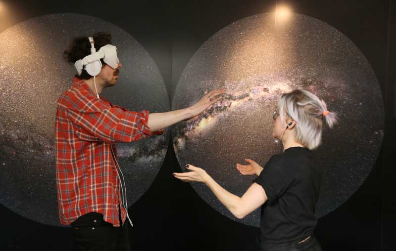 A blindfolded person in front of pictures of space with a smaller person guiding him