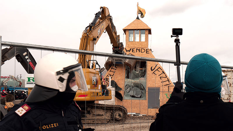 a teepee-like structure made for a protest is being taken down by a tractor with police in the foreground