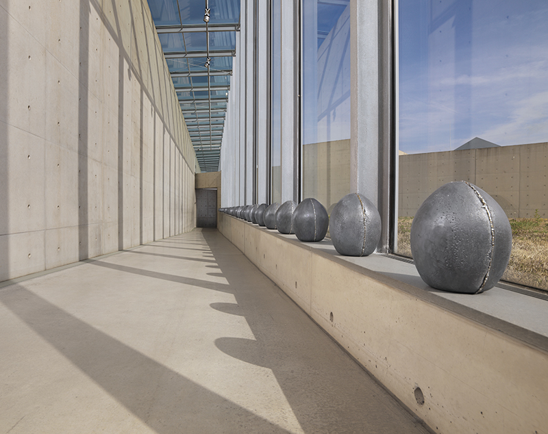 what look like large metal eggs are equally spaced apart on a window sill in a large museum with a glass facade