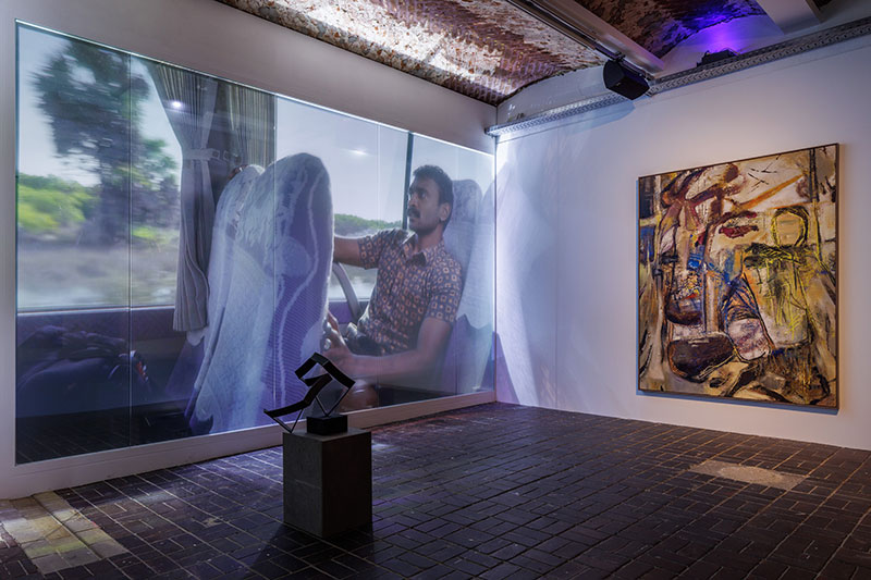 A photo of a gallery room with a projection on a wall and a painting hanging perpendicular to it