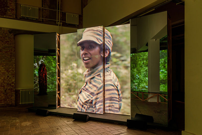 A photo of a video installation in a gallery consisting of multiple tall LED screens upon which a Sri Lankan woman can be seen