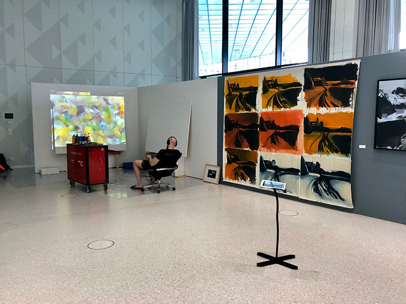 A man sitting in a large open space, what could be a gallery or studio, wearing headphones and looking at a projection on the wall