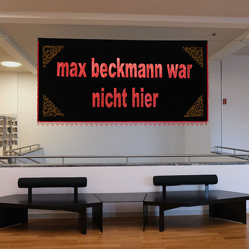 A banner hanging over an atrium in a public space upon which is embroidered "Max Beckmann war nicht hier"