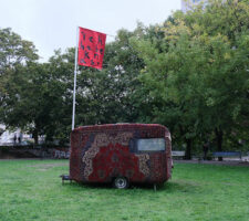 a camping trailer covered in persian rugs with a large red flag in a park-like setting