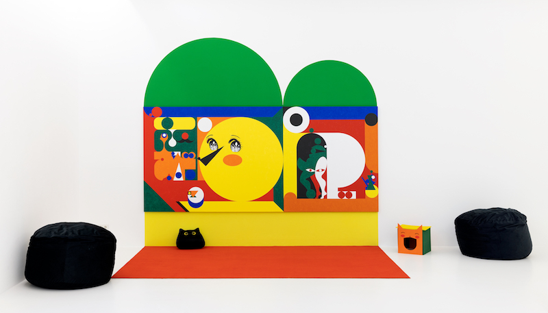 a vibrant wall installation in the white gallery space with bean bag chairs and cat houses on the floor