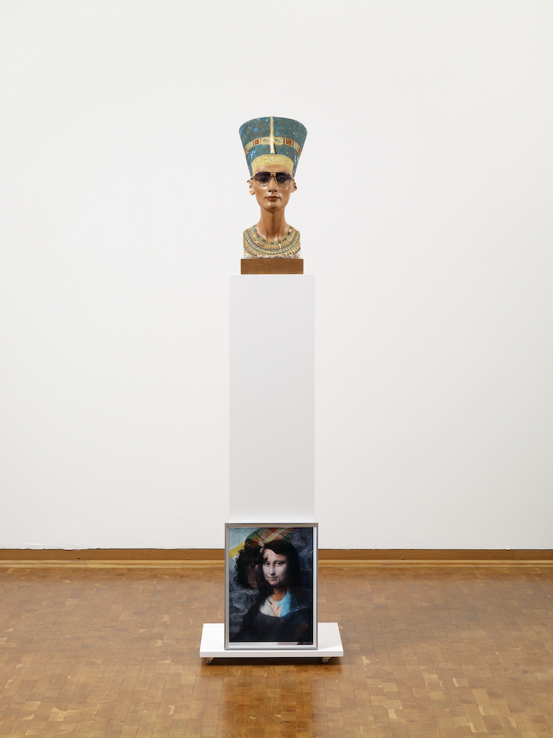 Nefertiti bust wearing sunglasses standing on a pedestal against which leans a photograph showing Mona Lisa