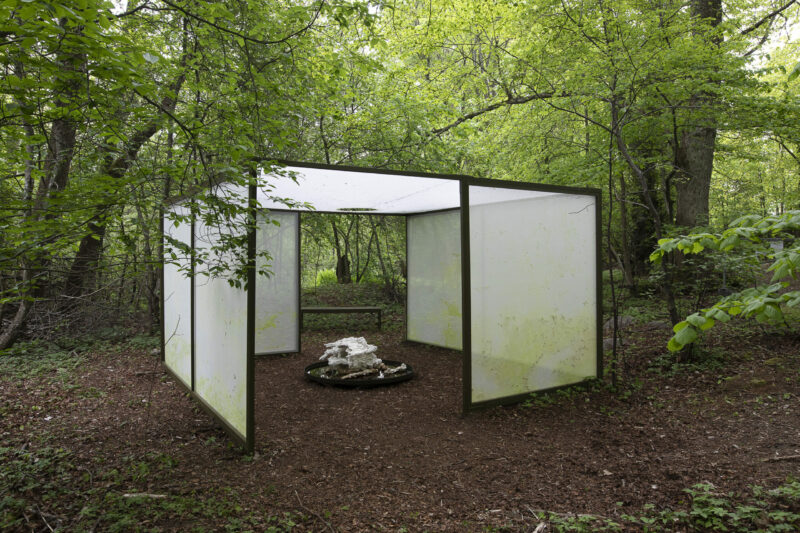 a square-shaped white cosntruction with fabric walls and a sculpture in the middle, placed in a forest