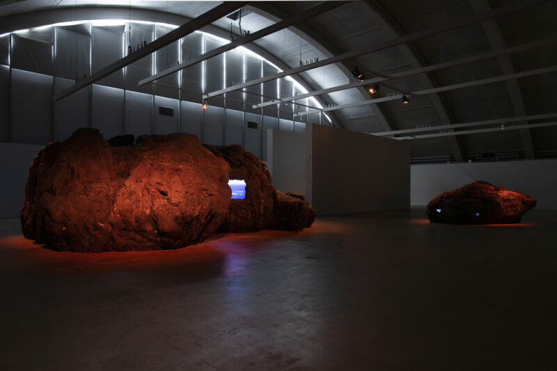 two large, rock-shaped sculptures with small screens embedded in them installed in a museum space