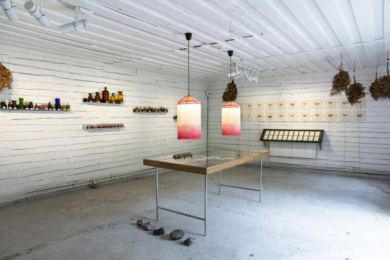 The inside of a white wooden shed with a table, two lamps, shelves on the walls storing bottles and drawings on the wall and in a vitrine