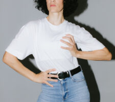 Portrait of Alexandra Bachzetsis wearing a white t-shirt and jeans, having one hand positioned on her breast and one on her hip