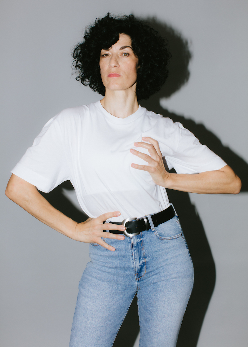 Portrait of Alexandra Bachzetsis wearing a white t-shirt and jeans, having one hand positioned on her breast and one on her hip